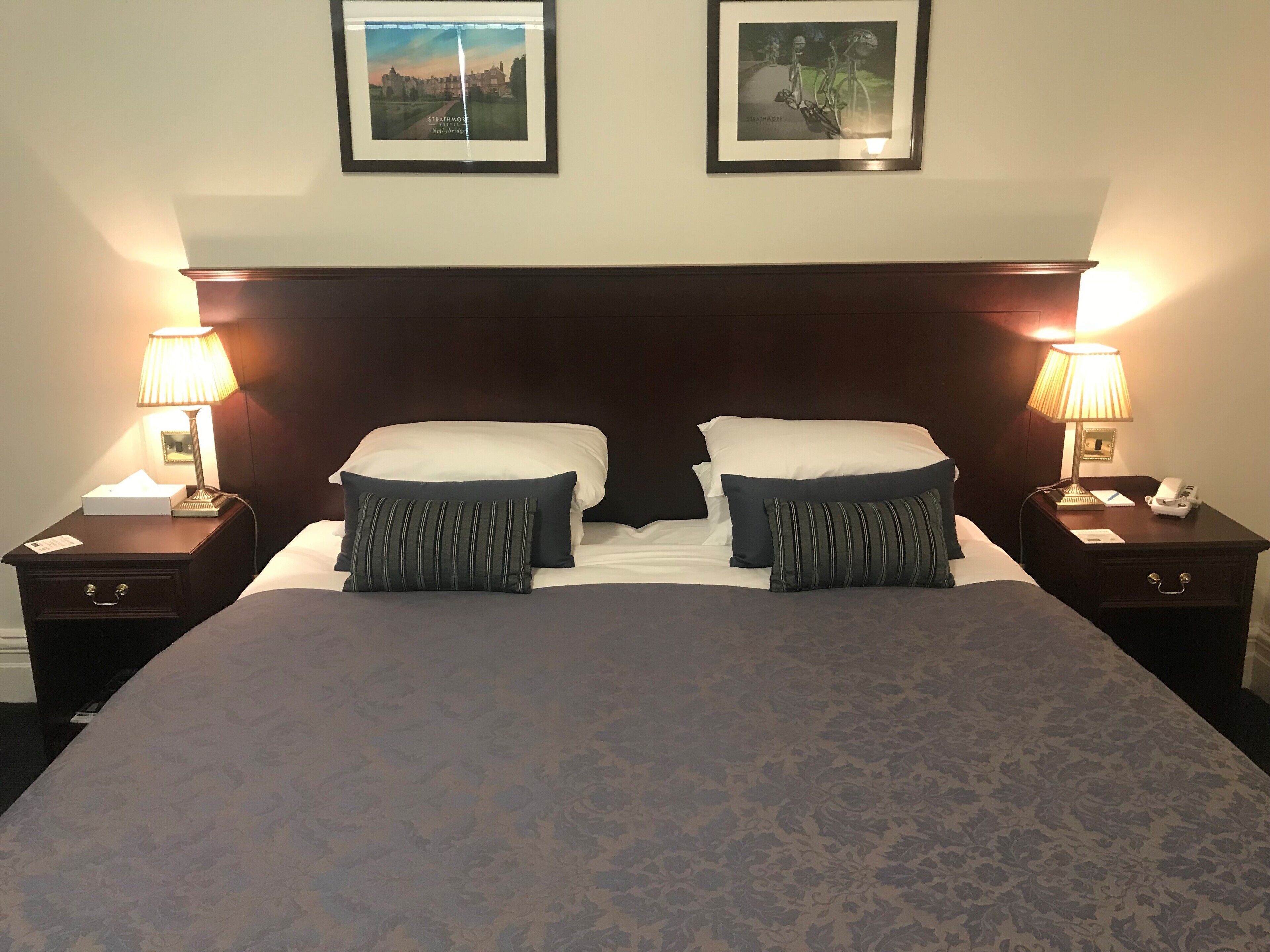 A neatly made double bed with two pillows, a nightstand with a lamp, and a framed picture above the bed.