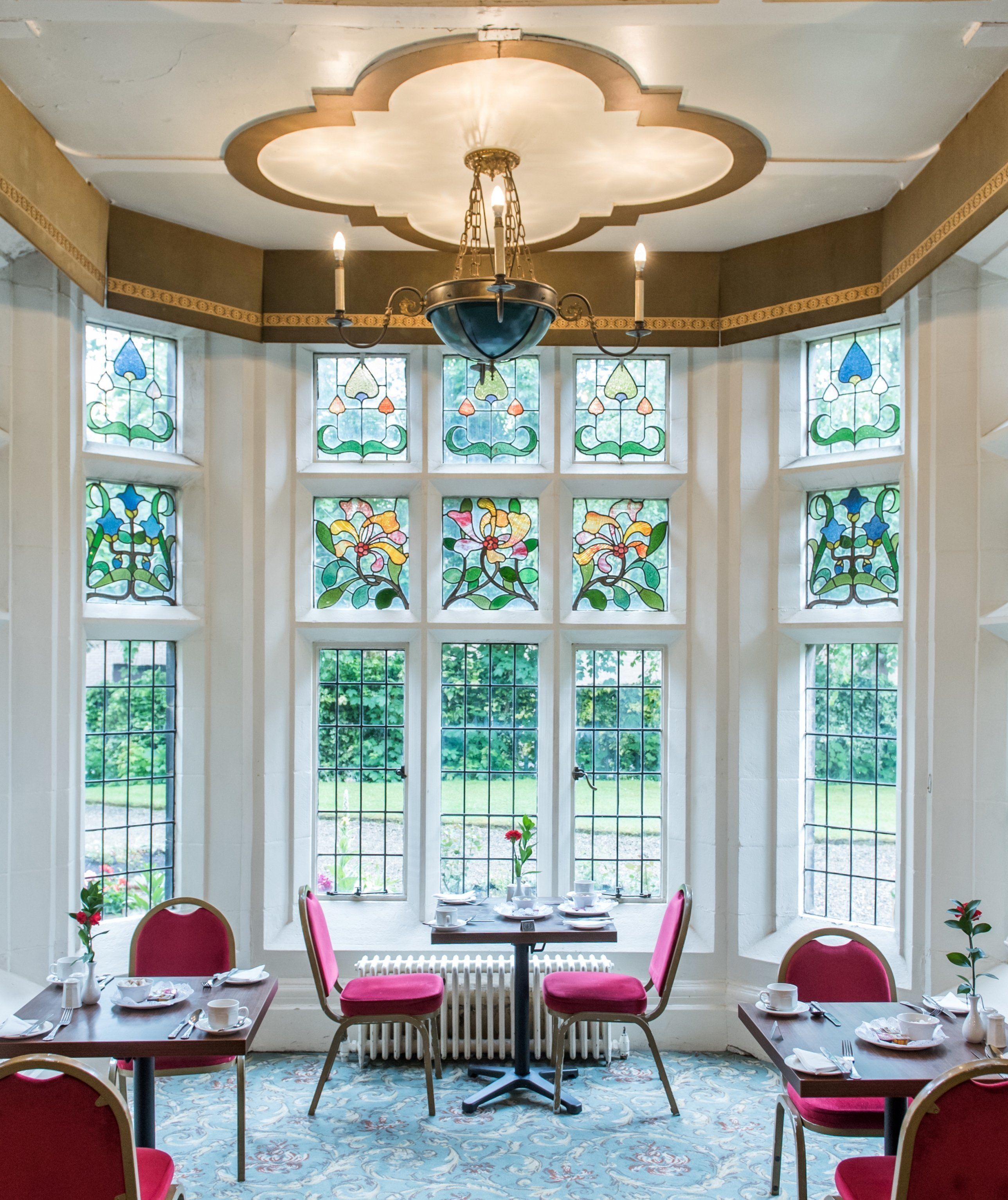 A dining area with a table set for two, surrounded by large stained glass windows, a chandelier, and a patterned carpet.