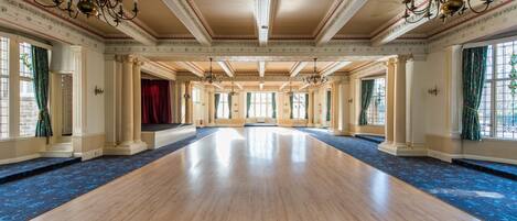 Un vaste hall doté d’un plancher en bois, de grandes fenêtres ornées de rideaux verts et de lustres décoratifs.