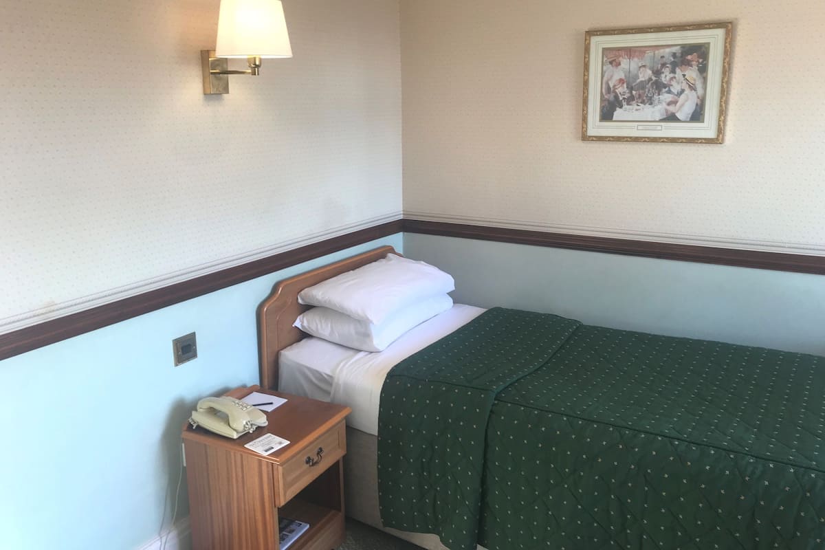 A hotel room with a single bed, a bedside table with a lamp, a telephone, and a framed picture on the wall.