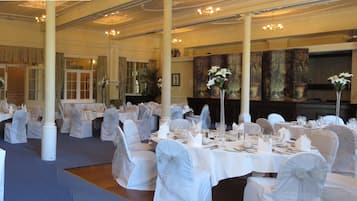 Een feestzaal met meerdere ronde tafels, elk gedekt met witte tafelkleden en stoelen, en een grote kroonluchter die aan het plafond hangt.