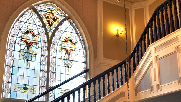 A grand staircase with a chandelier, ornate stained glass windows, and a polished wooden finish. - Cairn Hotel (Harrogate)