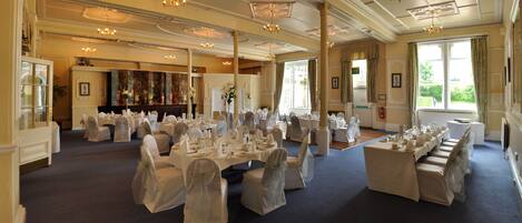 Un salón de banquetes con múltiples mesas redondas dispuestas para eventos, ventanas amplias y detalles arquitectónicos clásicos.