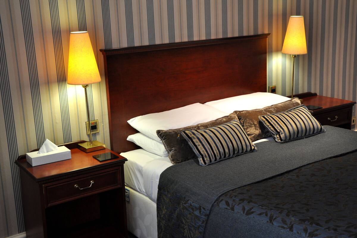 A hotel room with a wooden headboard, two bedside lamps, a bedside table with a tissue box and a phone, a bed with white linens and striped pillows, and striped wallpaper.