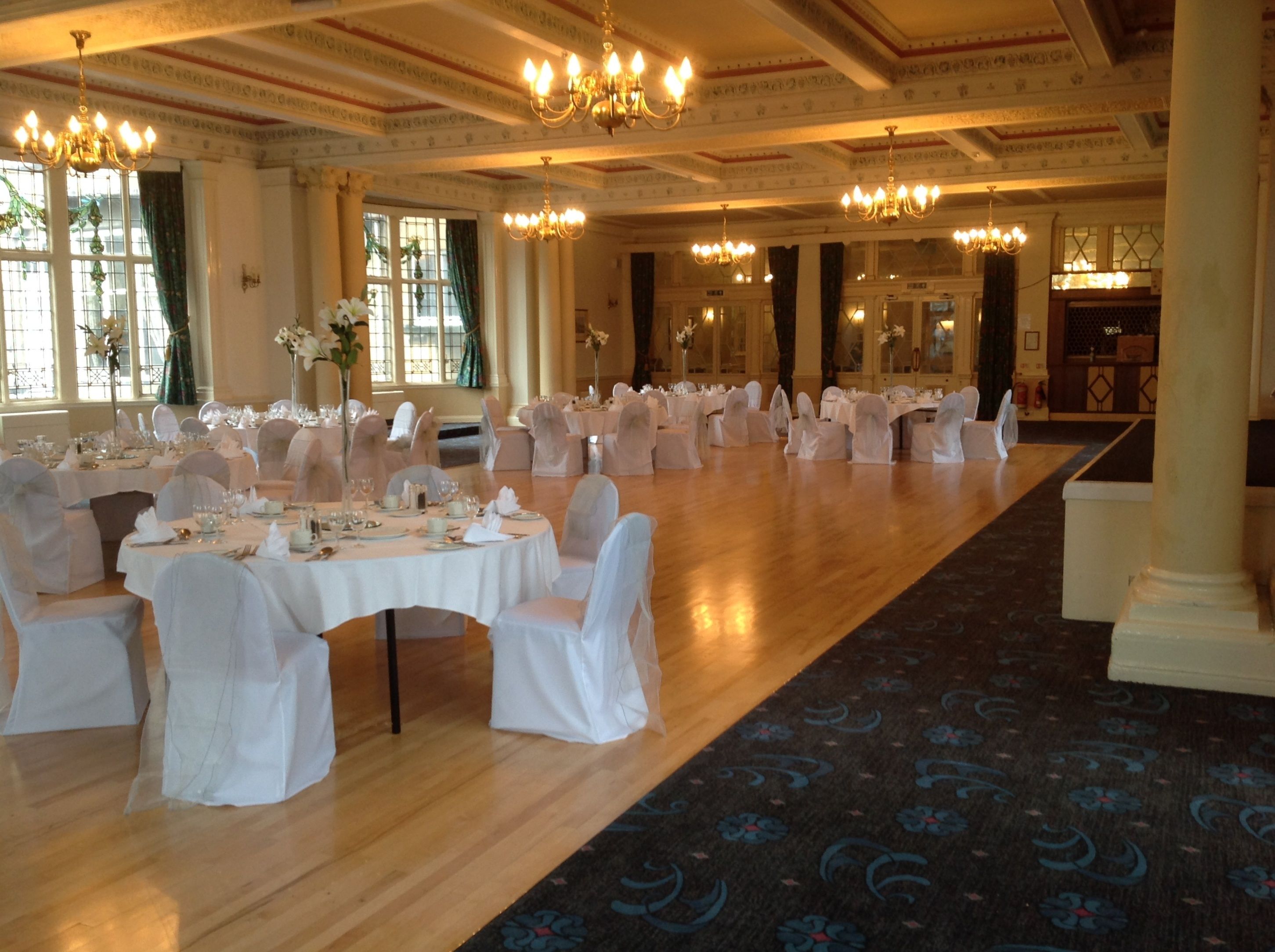 A banquet hall with round tables set for a formal event, featuring chandeliers and a patterned carpet.