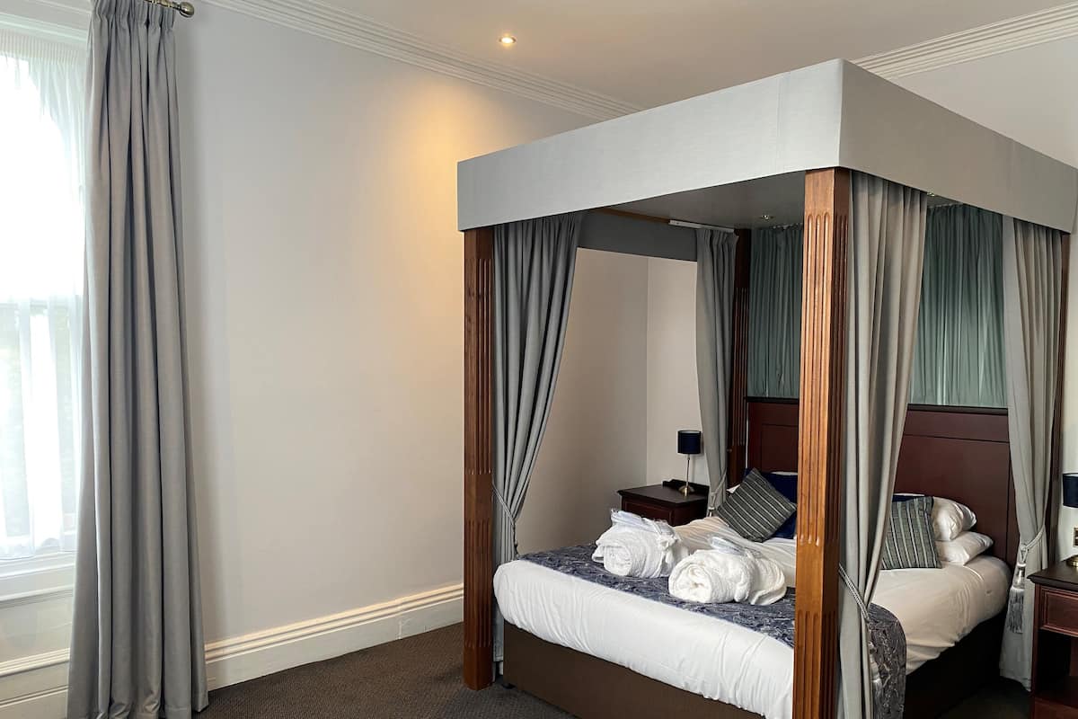 A four-poster canopy bed with a wooden headboard, a bedside table with a lamp, and a view of the outside through the window.