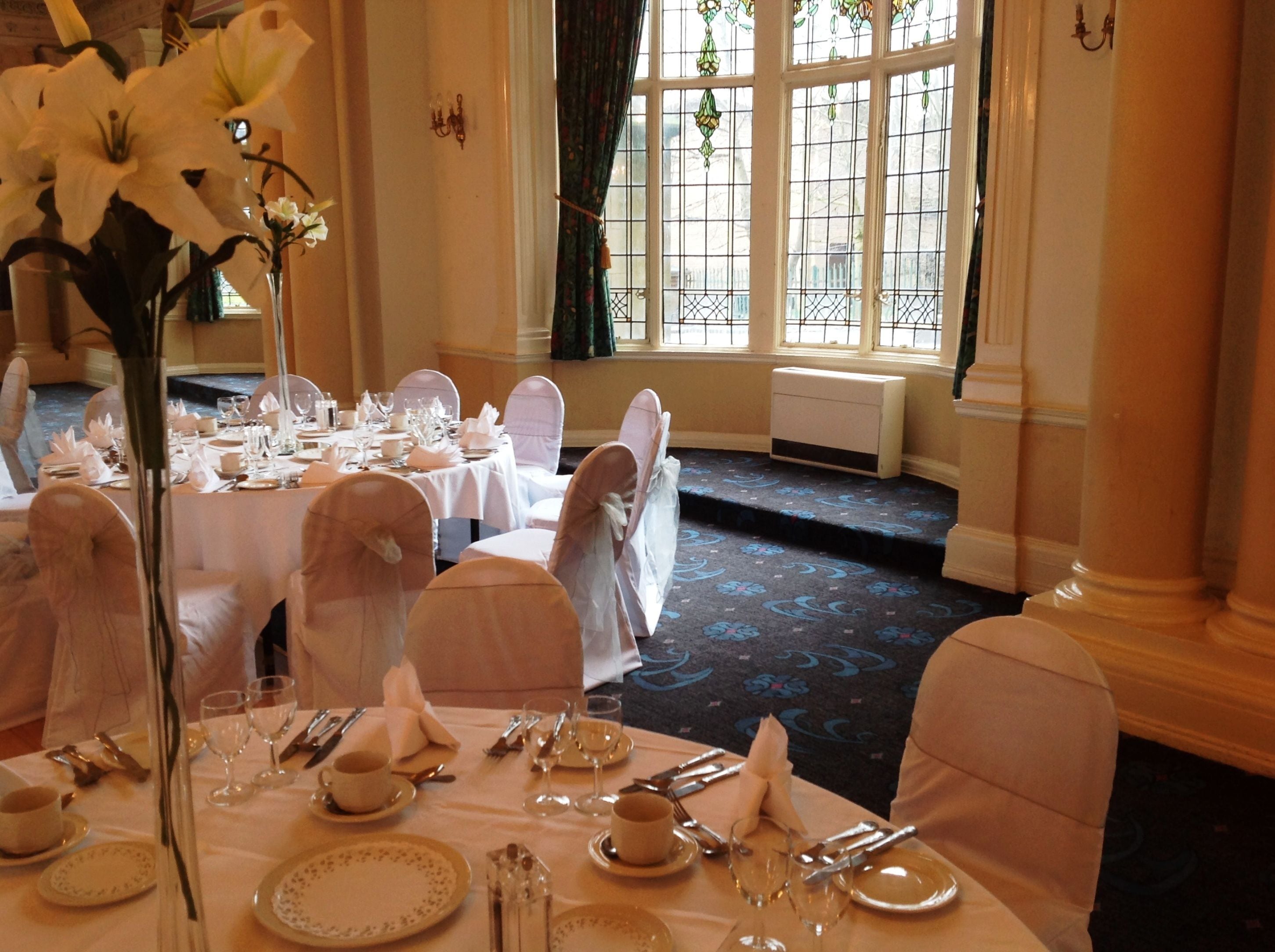 Une salle de banquet avec des tables rondes dressées pour un événement formel, agrémentée de nappes blanches, de chaises recouvertes de tissu blanc et de compositions florales en guise de centre de table.
