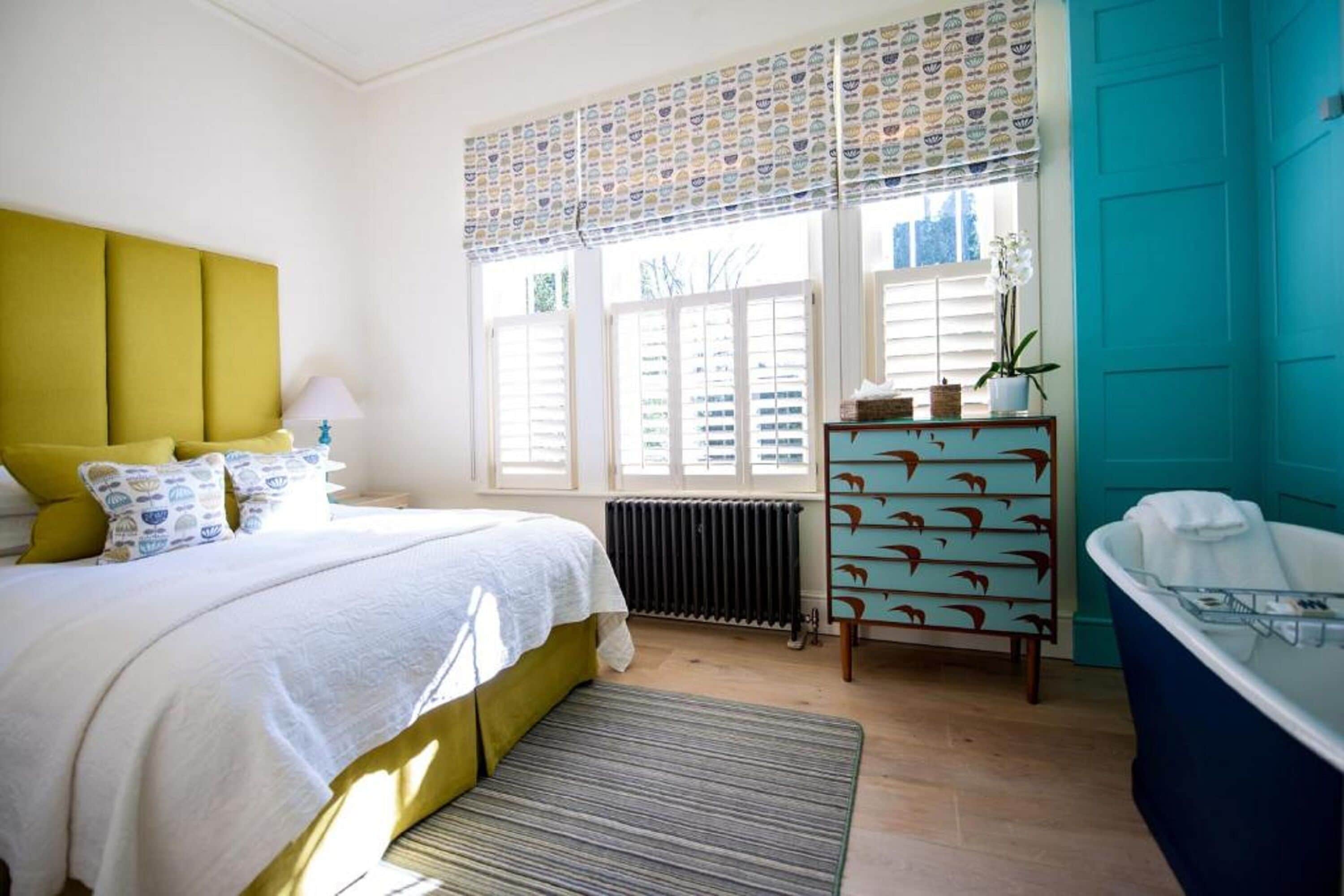 Une chambre à coucher comprenant un lit, une table de chevet, une commode, un radiateur, une fenêtre avec des stores et une baignoire.