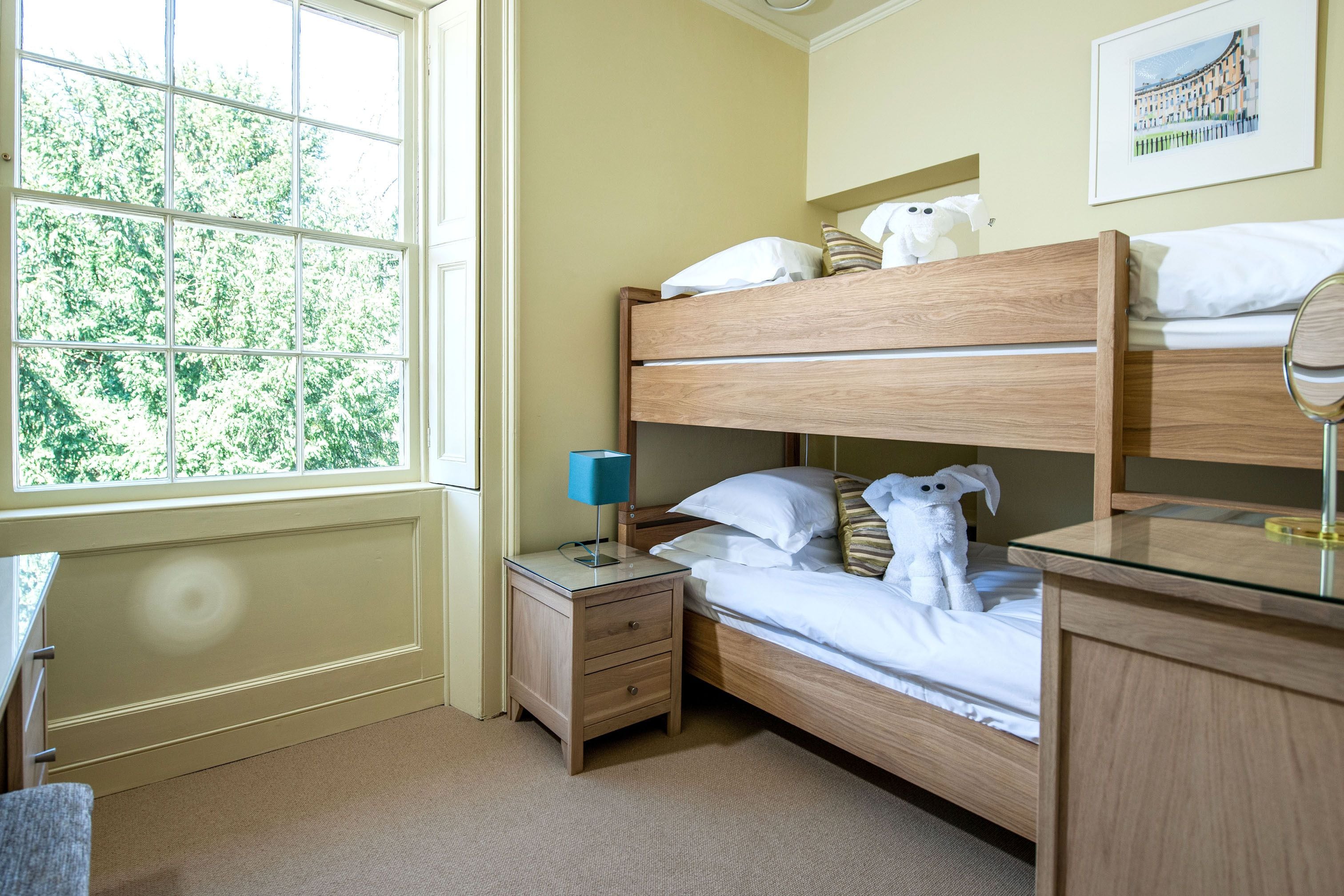 A room with a bunk bed, a bedside table with a lamp, a small drawer, and a window with a view of trees.