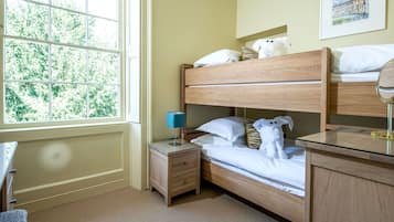 A room with a bunk bed, a bedside table with a lamp, a small drawer, and a window with a view of trees.