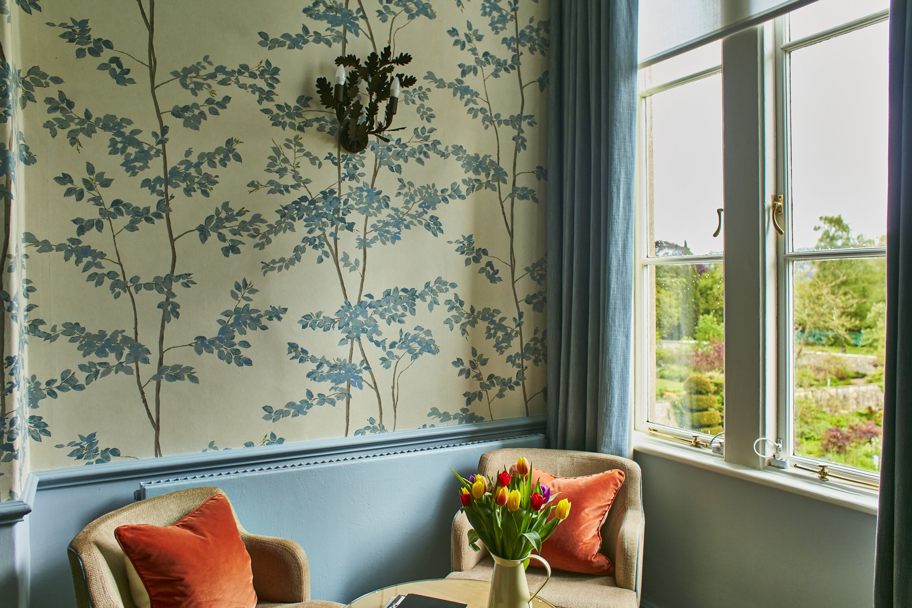 Quarto com papel de parede floral azul e branco, janela com vista para vegetação e um vaso com flores coloridas sobre uma mesa.