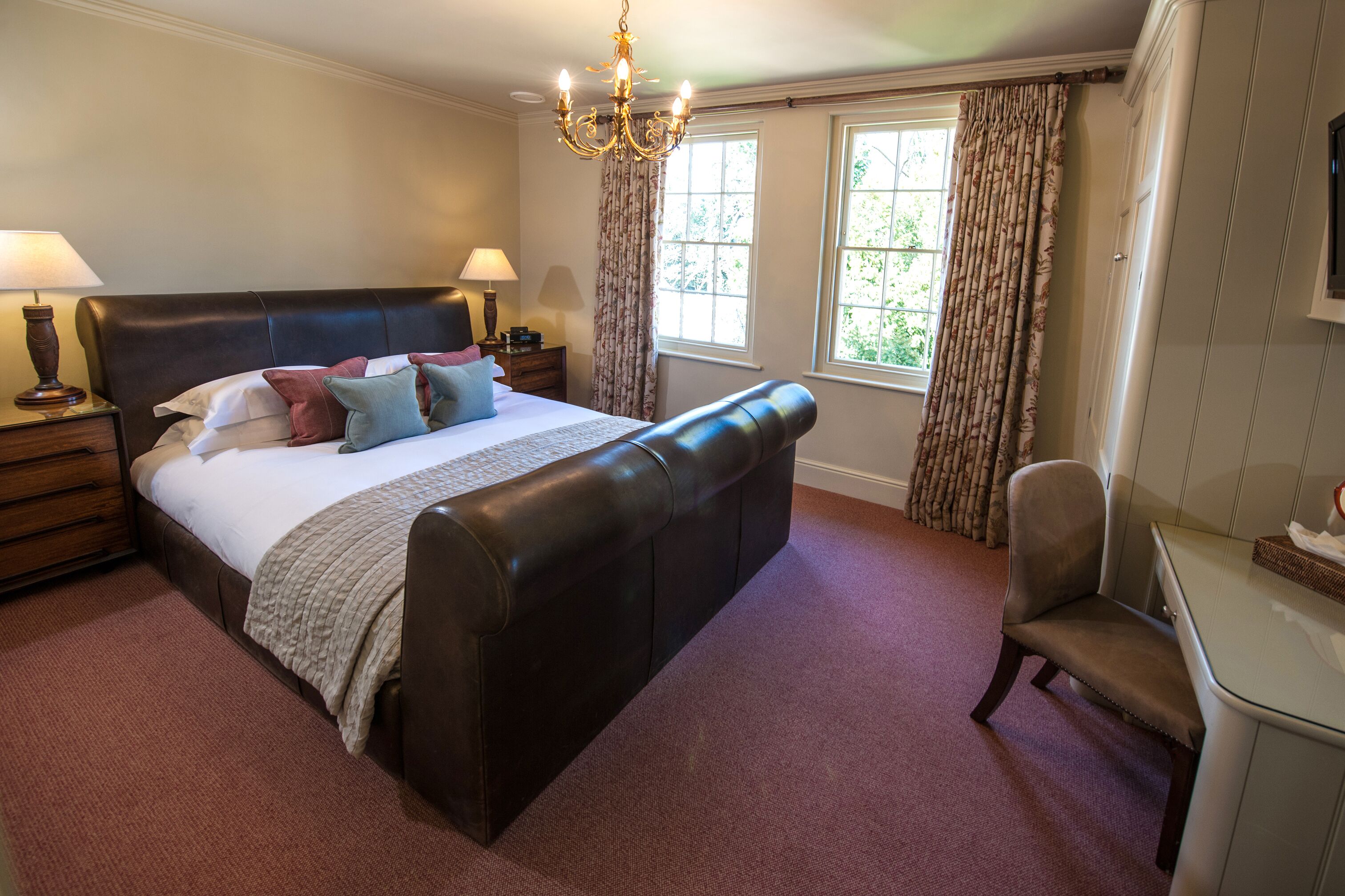 A bedroom with a large bed, two bedside tables with lamps, a chair, a desk, and a window with curtains.