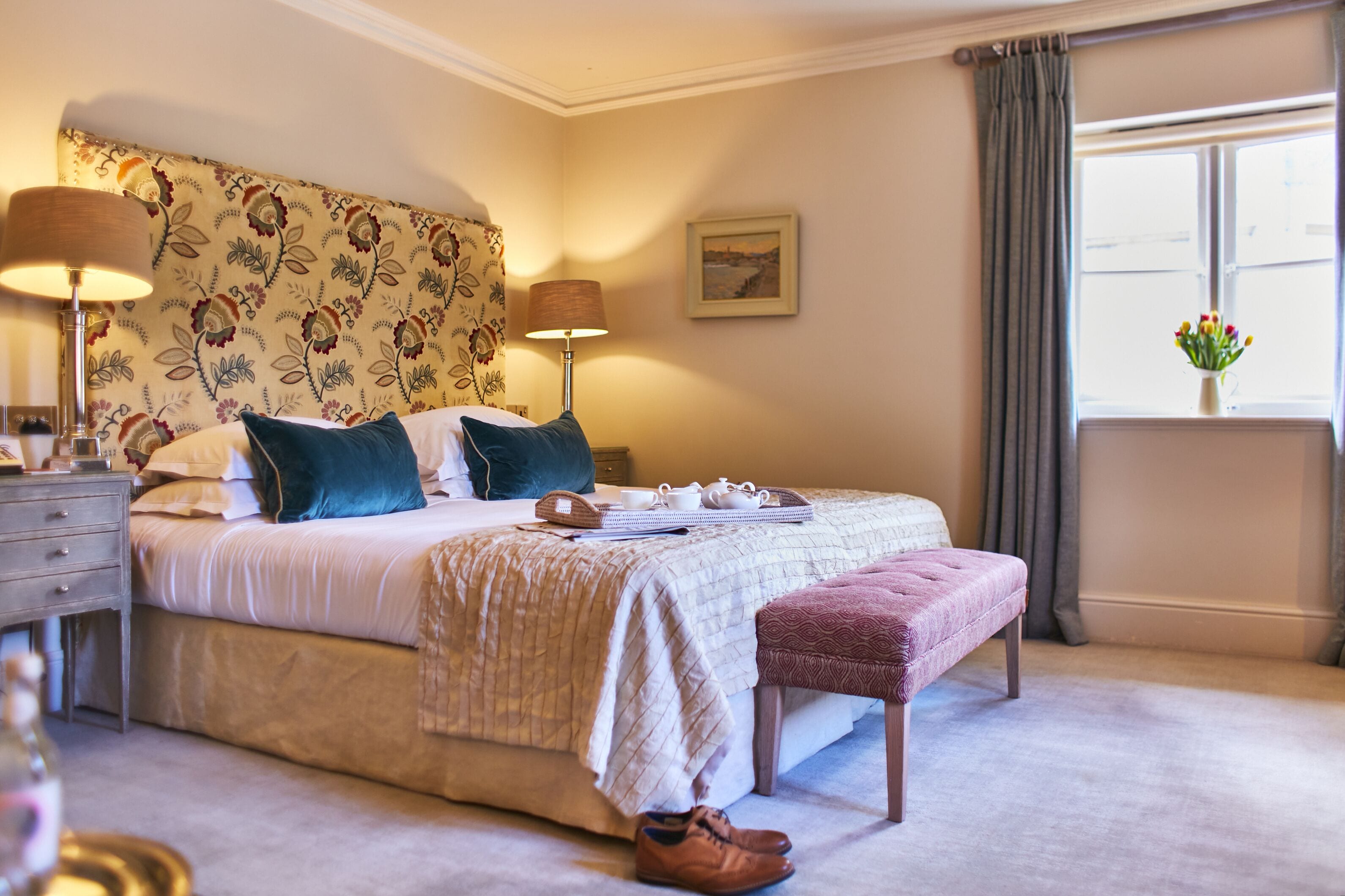 Un dormitorio ordenado con cama, lámparas de noche, mesitas de noche, una ventana con cortinas y un jarrón con flores.