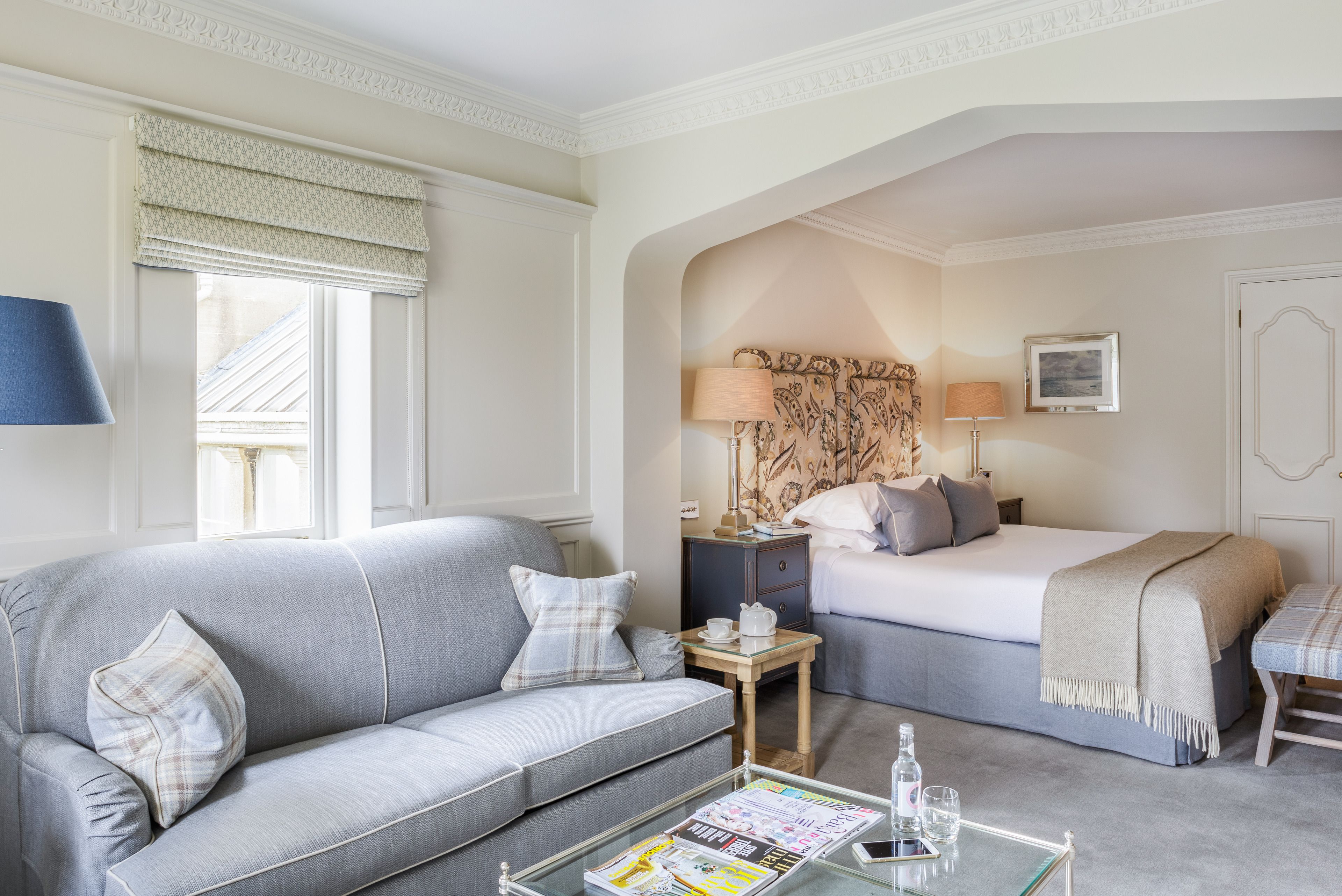 A hotel room with a grey sofa, a bed with a headboard, a nightstand, a table with a magazine, and a window with a curtain.