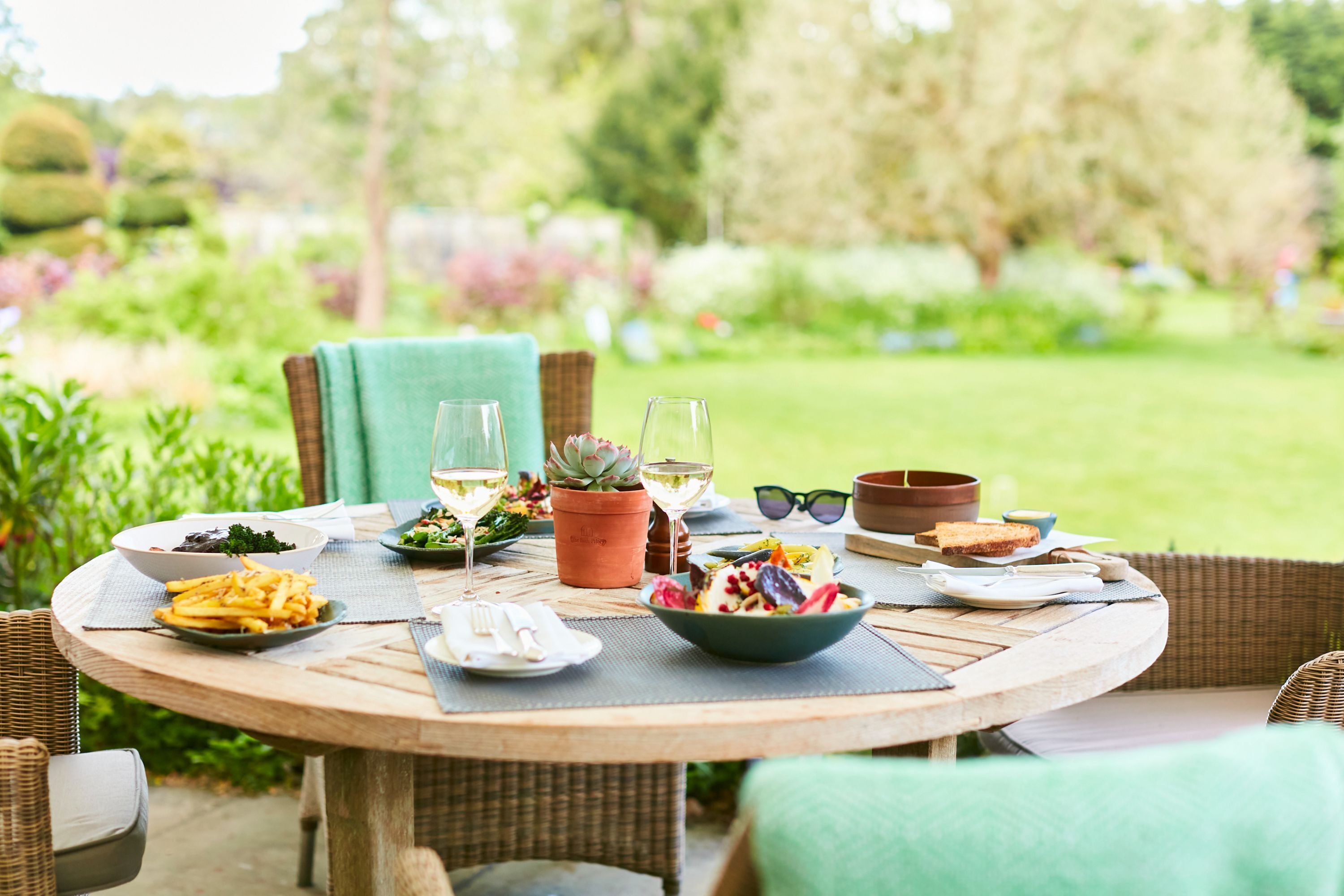 A round wooden table set outdoors with various dishes, glasses, and a potted plant.