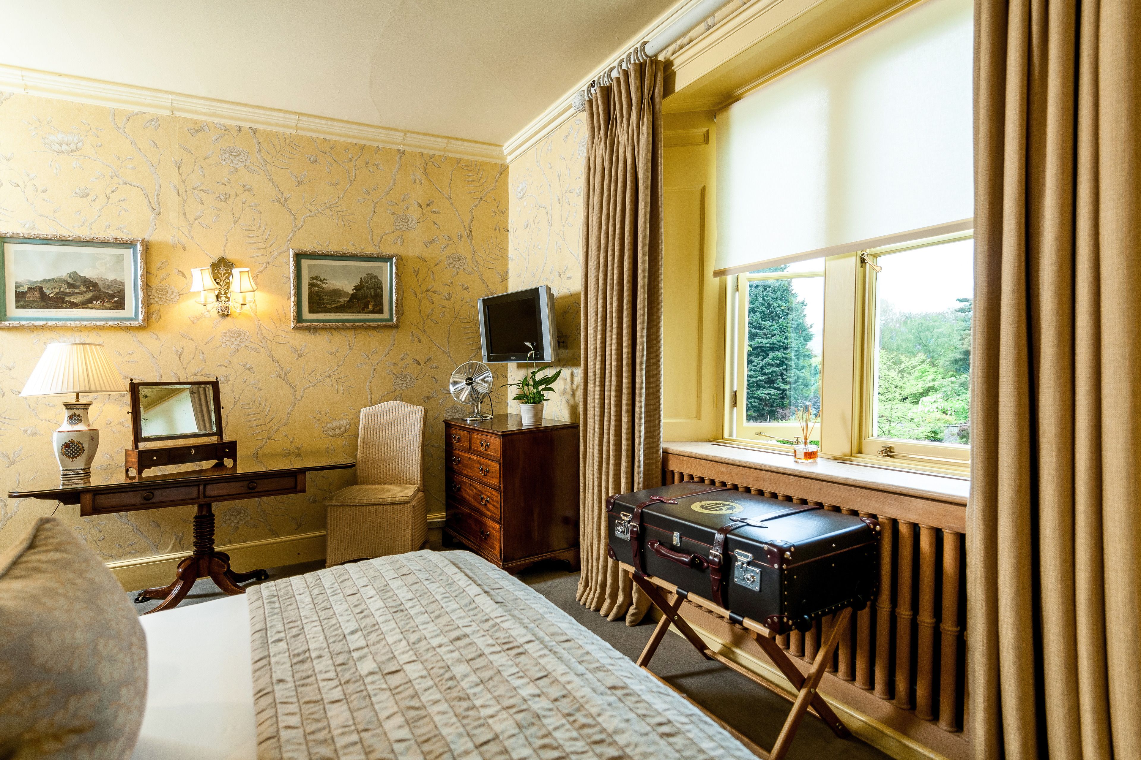 Ein Hotelzimmer mit Bett, Schreibtisch mit Lampe, Kommode, Fernseher und Koffer.