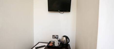 A hotel room with a flat-screen TV mounted on the wall, a small table with a remote control, a coffee maker, and a magazine.