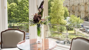 Een kamer met een ronde glazen tafel, twee fauteuils, een vaas met bloemen en uitzicht op een boom en gebouwen door grote ramen.