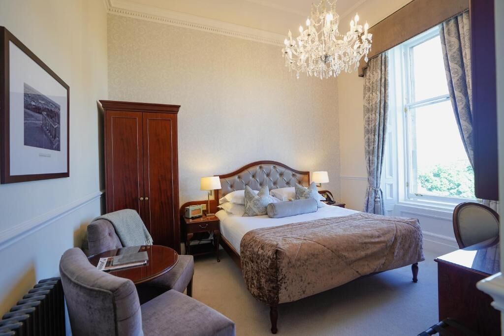 Een hotelkamer met een groot bed, een zithoek met een stoel en een tafeltje, een houten garderobe en een kroonluchter.