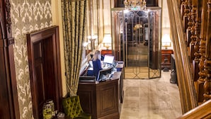 Een statige lobby met een houten trap, een bureau met een computer en een groene fauteuil.