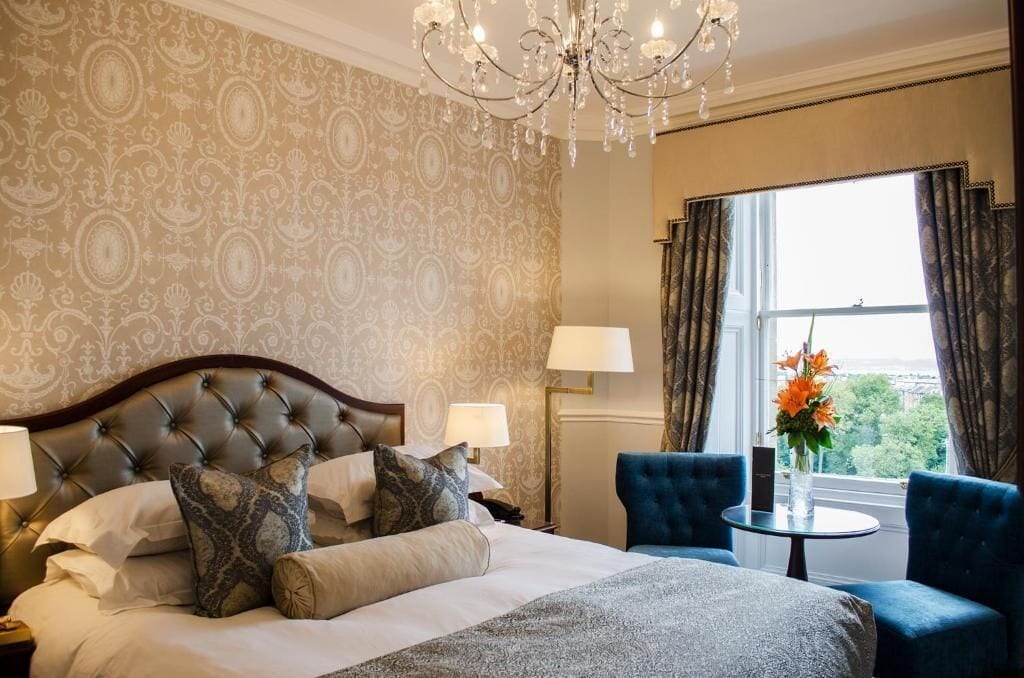 Hotelkamer met een gestoffeerd hoofdeinde, een bed met gekleurde kussens, een ronde tafel met een bloevas, en uitzicht op het omliggende landschap door een raam met gordijnen.