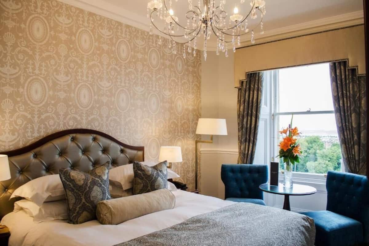Hotelkamer met een gestoffeerd hoofdeinde, een bed met gekleurde kussens, een ronde tafel met een bloevas, en uitzicht op het omliggende landschap door een raam met gordijnen.