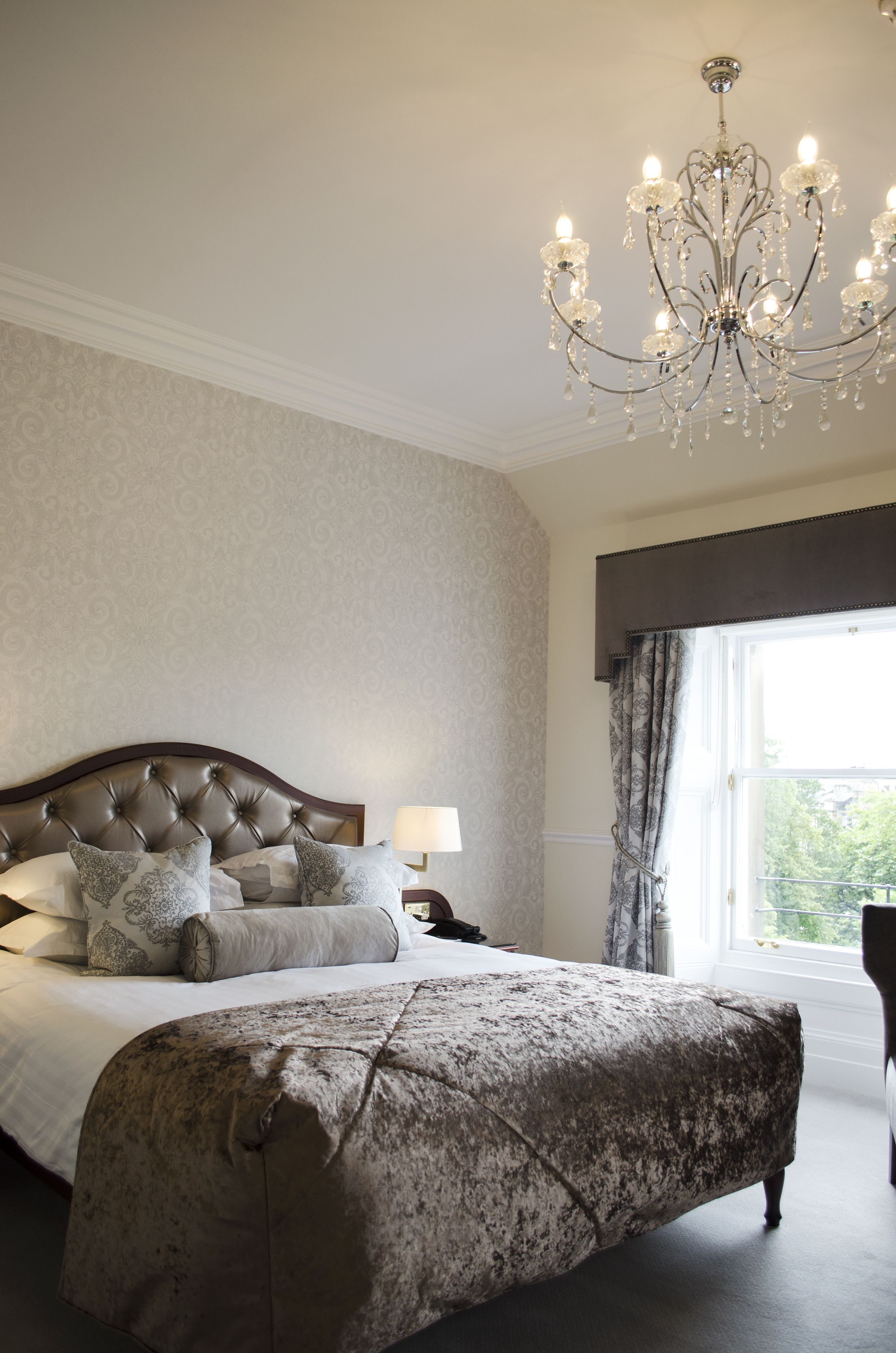 A bedroom with a tufted headboard, a large bed with multiple pillows, a bench, a chandelier, and a window with curtains.