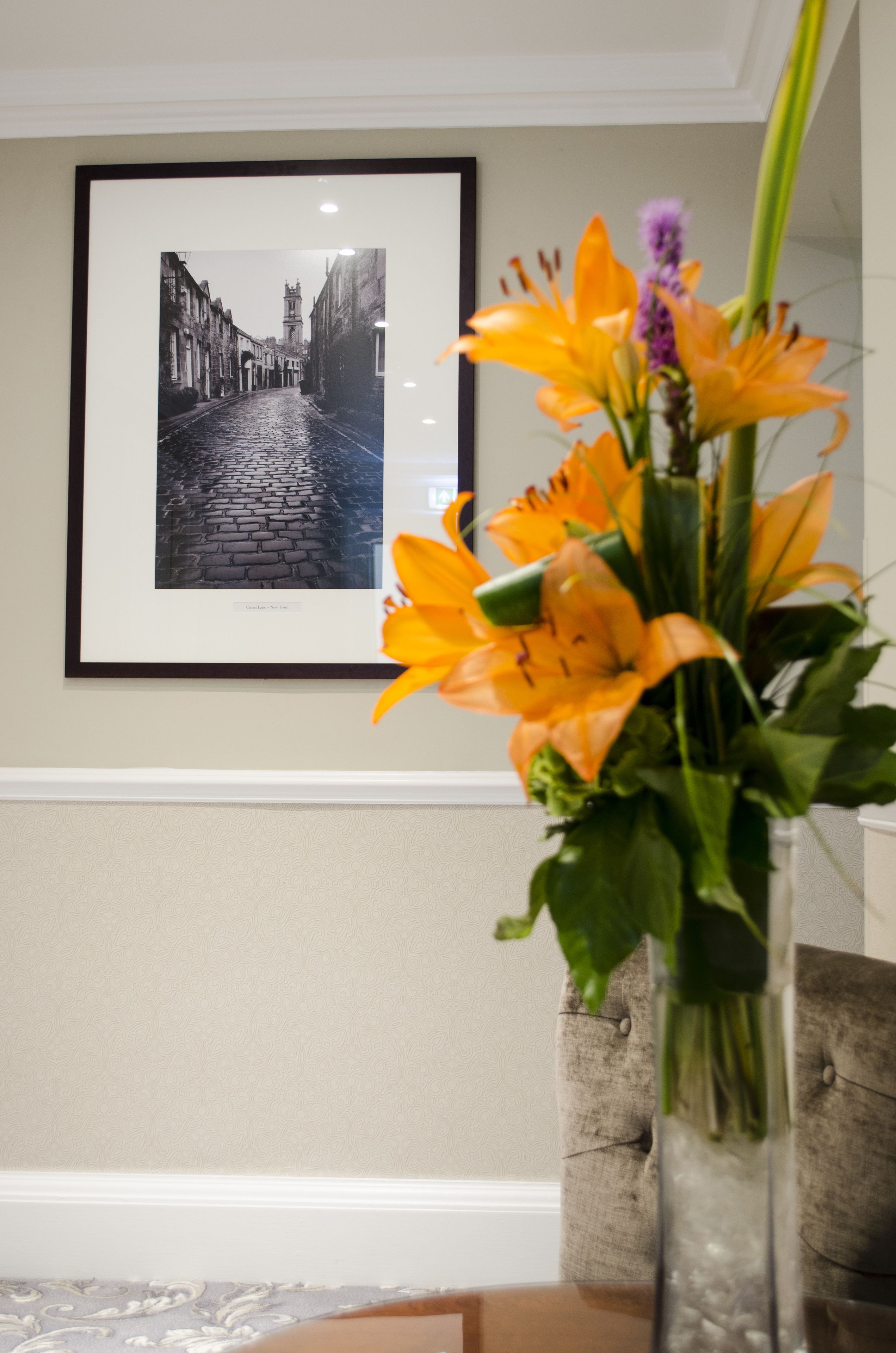 A framed photograph of a cobblestone street on a wall, a vase with orange flowers in the foreground, and a tufted chair in the background.