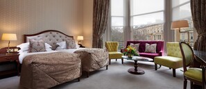 A hotel room with a large bed, two armchairs, a small table, and a view of buildings through large windows.