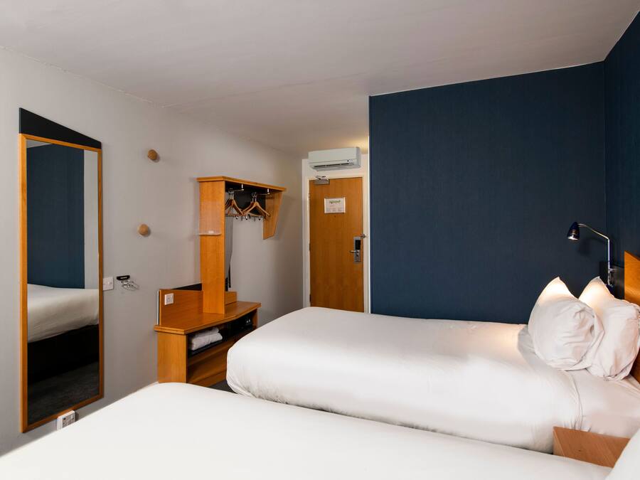 A hotel room with two beds, a wooden dresser, a mirror, a coat rack, and a nightstand with a lamp.