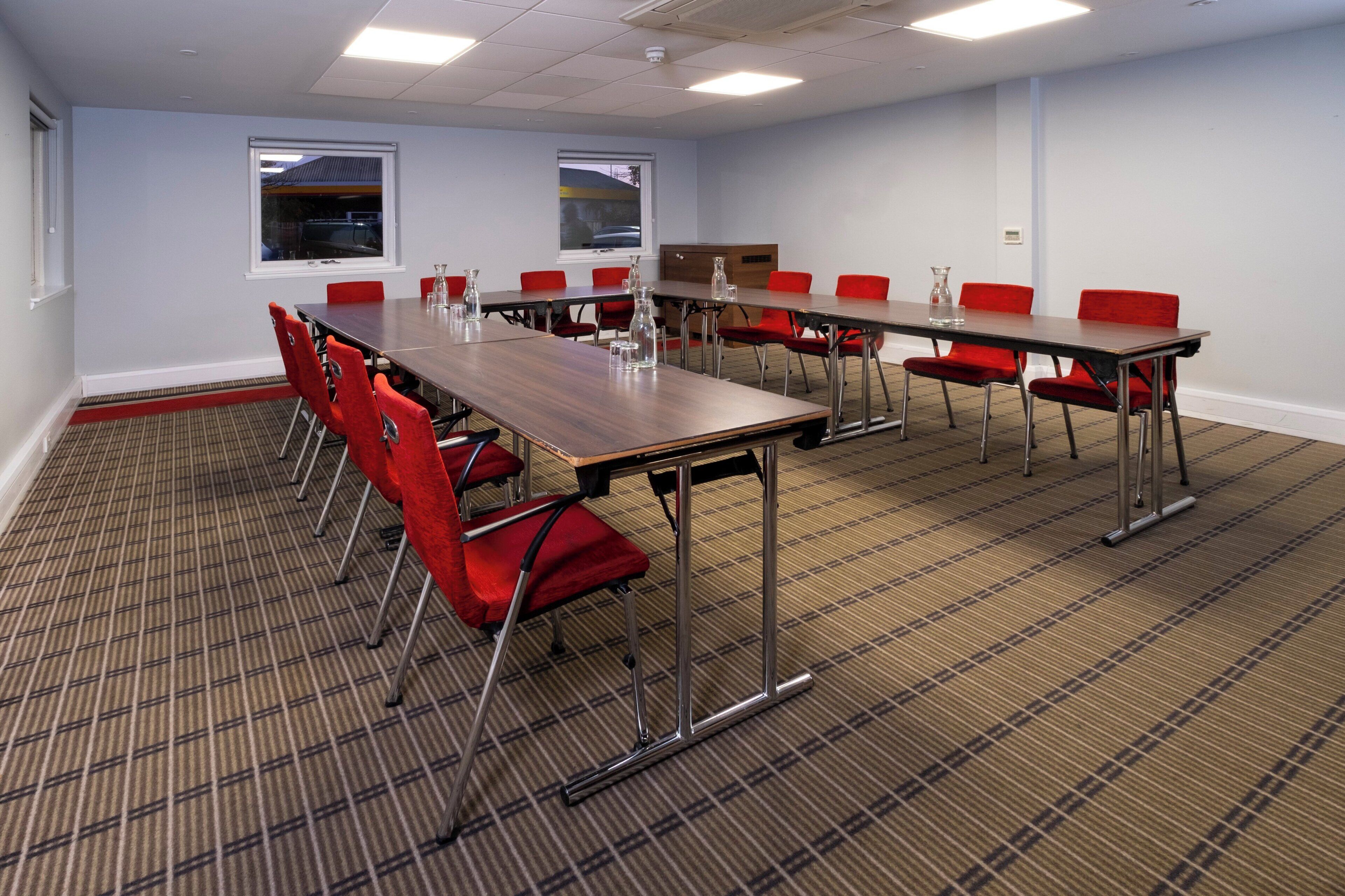A conference room with a long table, red chairs, and two windows.