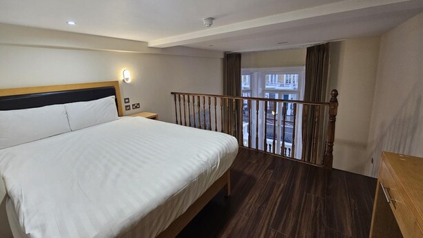 A hotel room with a large bed, a nightstand, a wooden railing, and a view of the street through the window. - Cromwell International Hotel (London)