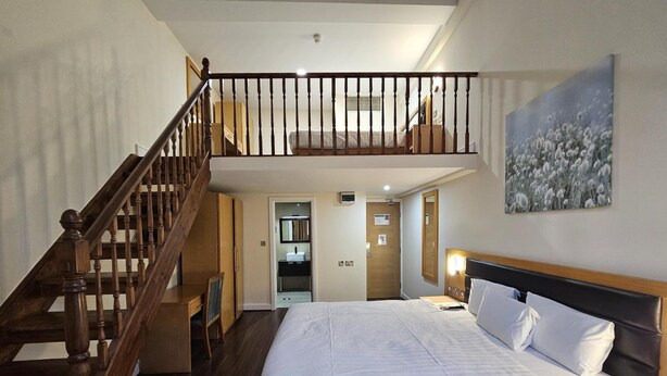 A bedroom with a wooden staircase, a bed with white bedding, a nightstand, a painting on the wall, and a bathroom visible through an open door. - Cromwell International Hotel (London)