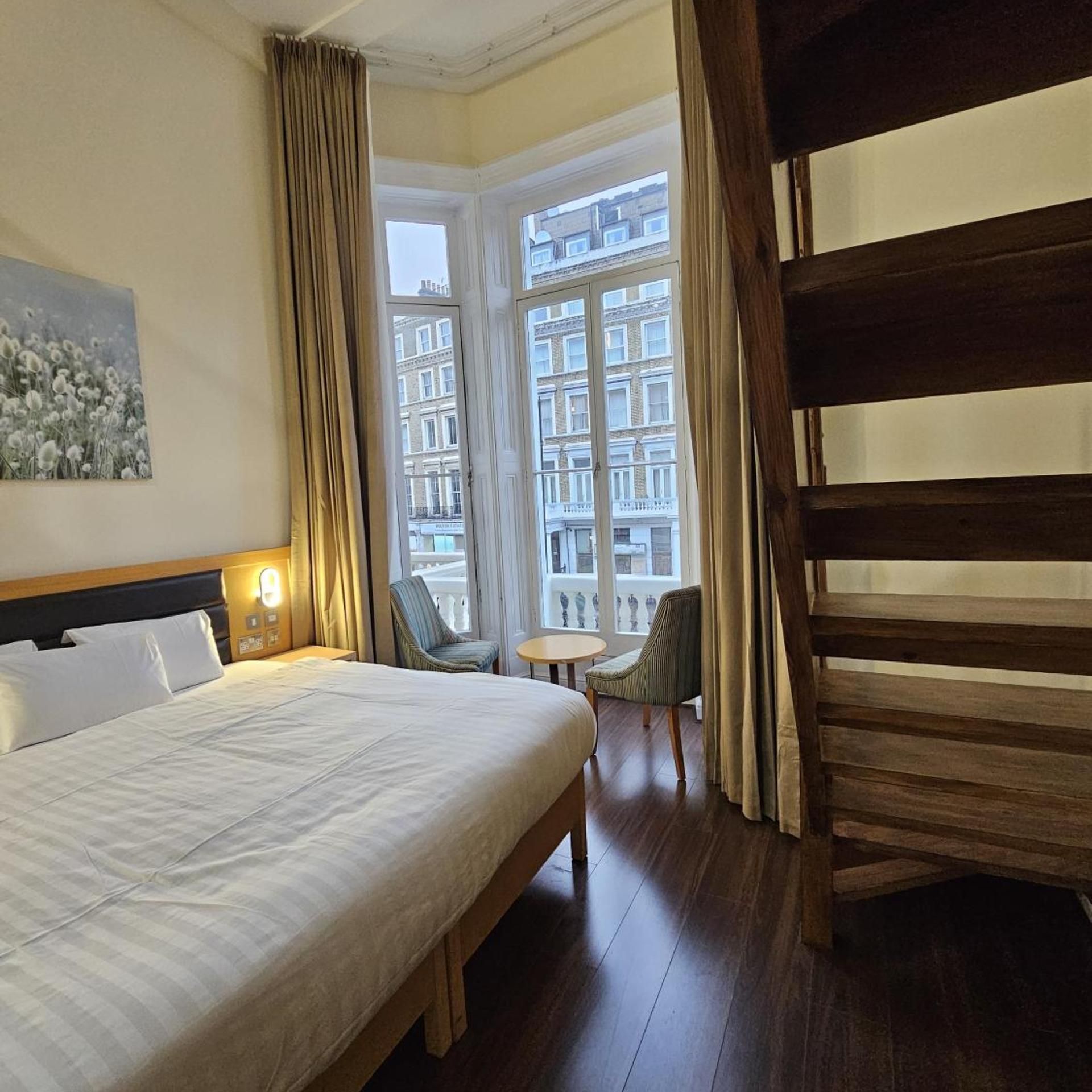 Habitación de hotel con una cama grande, una mesita, una silla y vistas a edificios por la ventana.