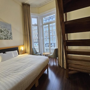 A hotel room with a large bed, a small table, a chair, and a view of buildings through the window. - Cromwell International Hotel (London)