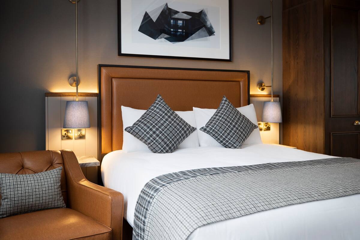 Quarto de hotel com uma cama grande, duas luminárias de cabeceira, um encosto de cama, uma poltrona de couro marrom e um quadro na parede.