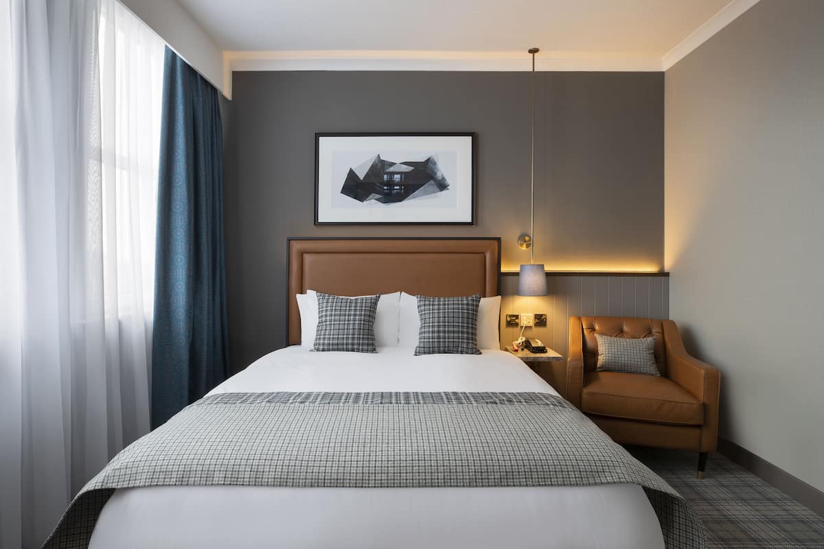 Quarto de hotel com uma cama grande, cabeceira, mesa de apoio com abajur, quadro decorativo na parede e janela com cortinas.