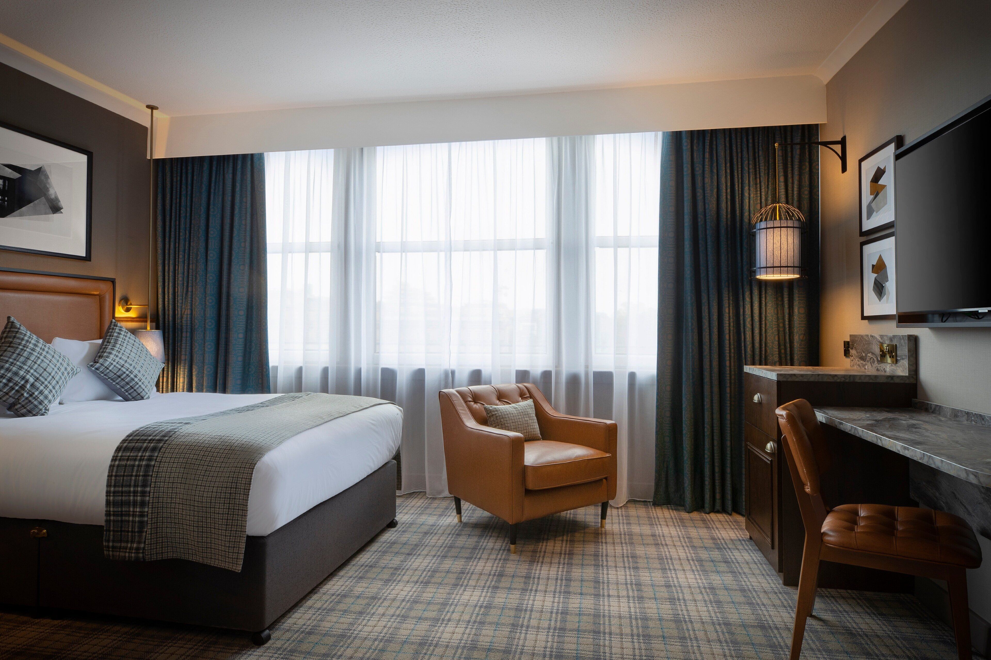 A hotel room with a bed, a chair, a desk, and a large window with curtains.