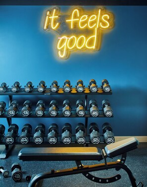 A gym with a neon sign that reads "it feels good" and a rack of dumbbells.