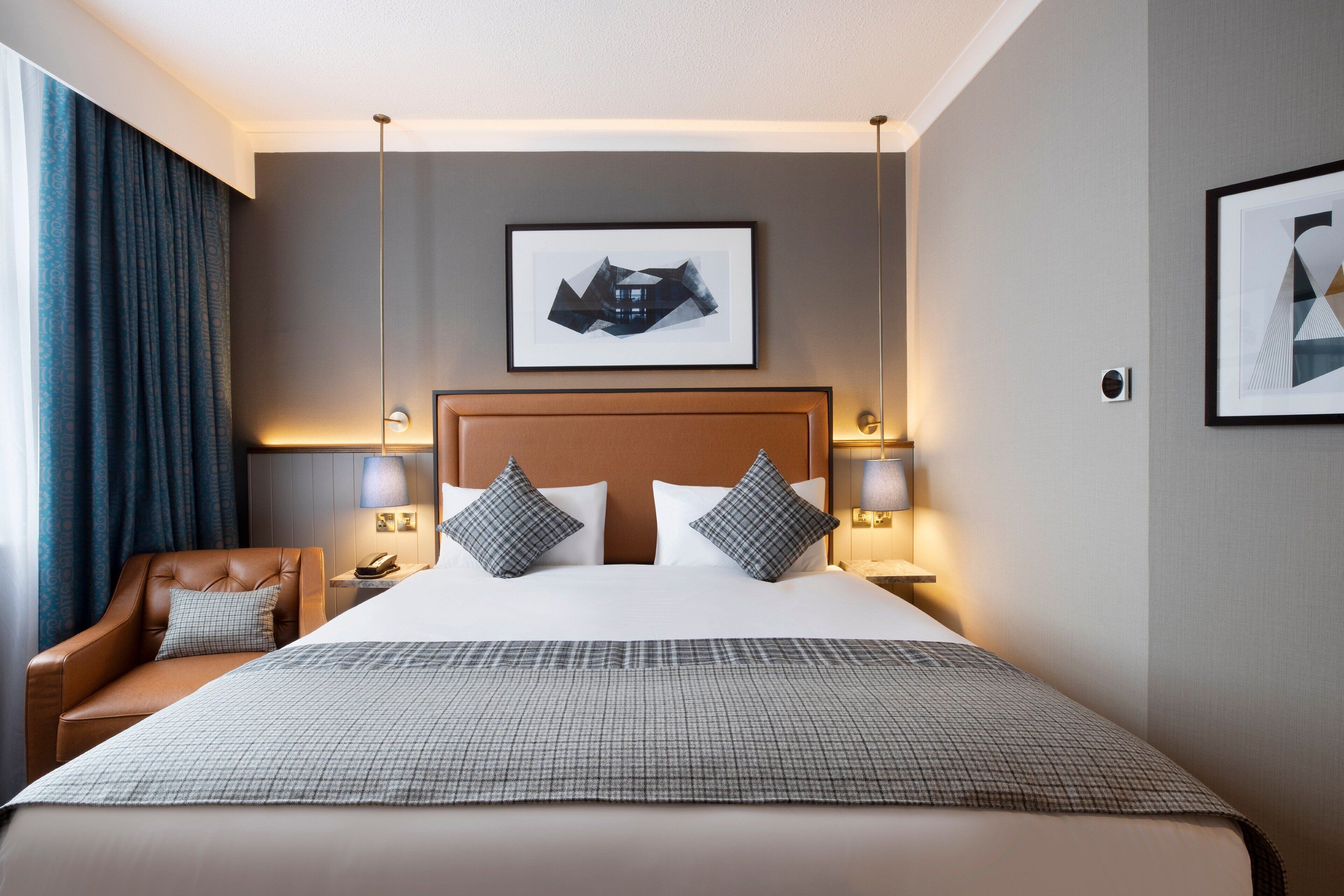 Hotelkamer met een groot bed, twee bedlampen, een hoofdeinde, een lijst met een afbeelding aan de muur, een stoel en een raam met gordijnen.