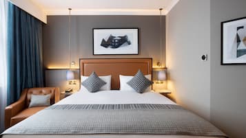 Hotelkamer met een groot bed, twee bedlampen, een hoofdeinde, een lijst met een afbeelding aan de muur, een stoel en een raam met gordijnen.