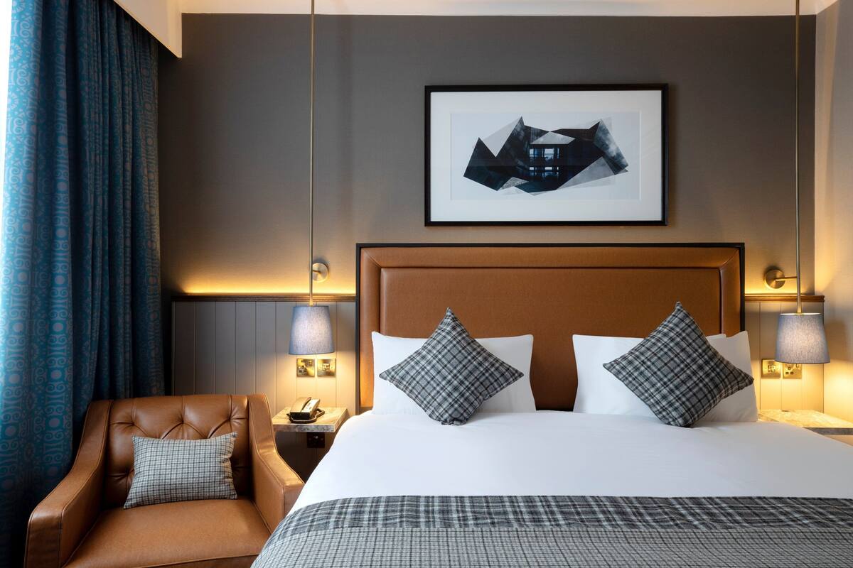 Una habitación de hotel con una cama grande, dos lámparas de noche, un sillón de cuero marrón y un cuadro en la pared.