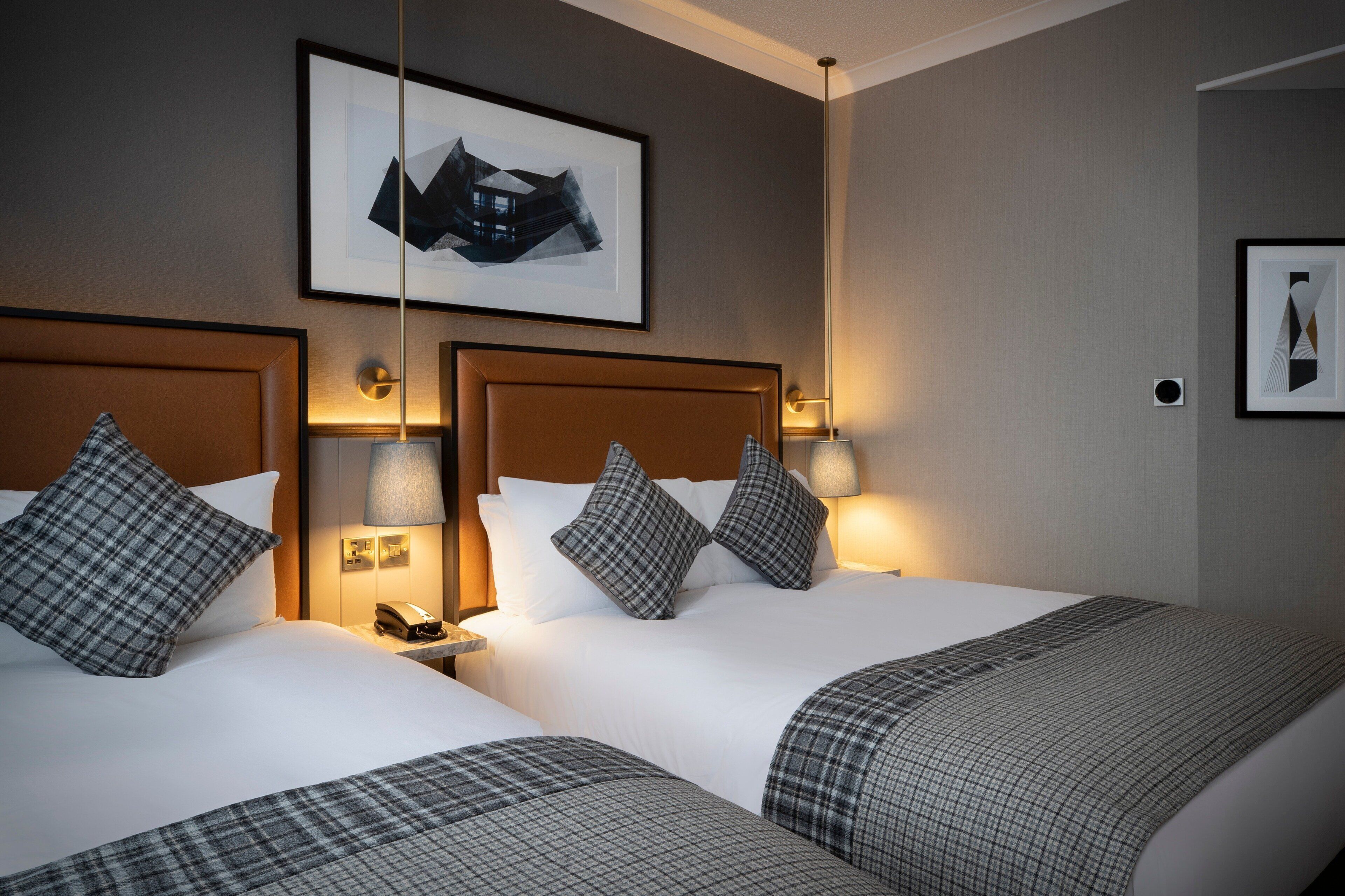 Ein Hotelzimmer mit zwei Betten, jeweils mit Kopfteil, Nachttischlampen und Telefon. Über den Betten hängt ein gerahmtes Bild an der Wand.