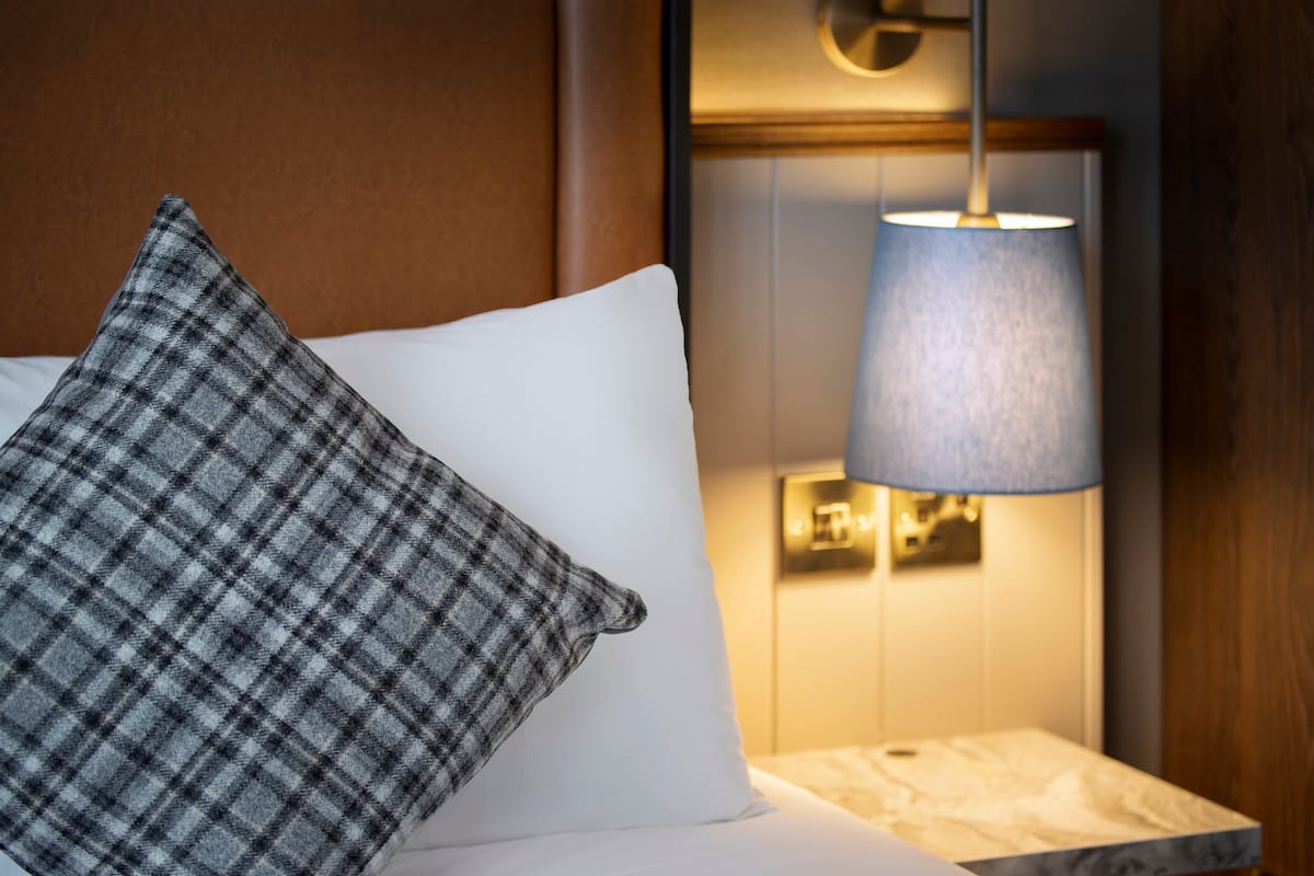 Quarto de hotel com cama, almofada xadrez, almofada branca, mesa de cabeceira e luminária fixada na parede.
