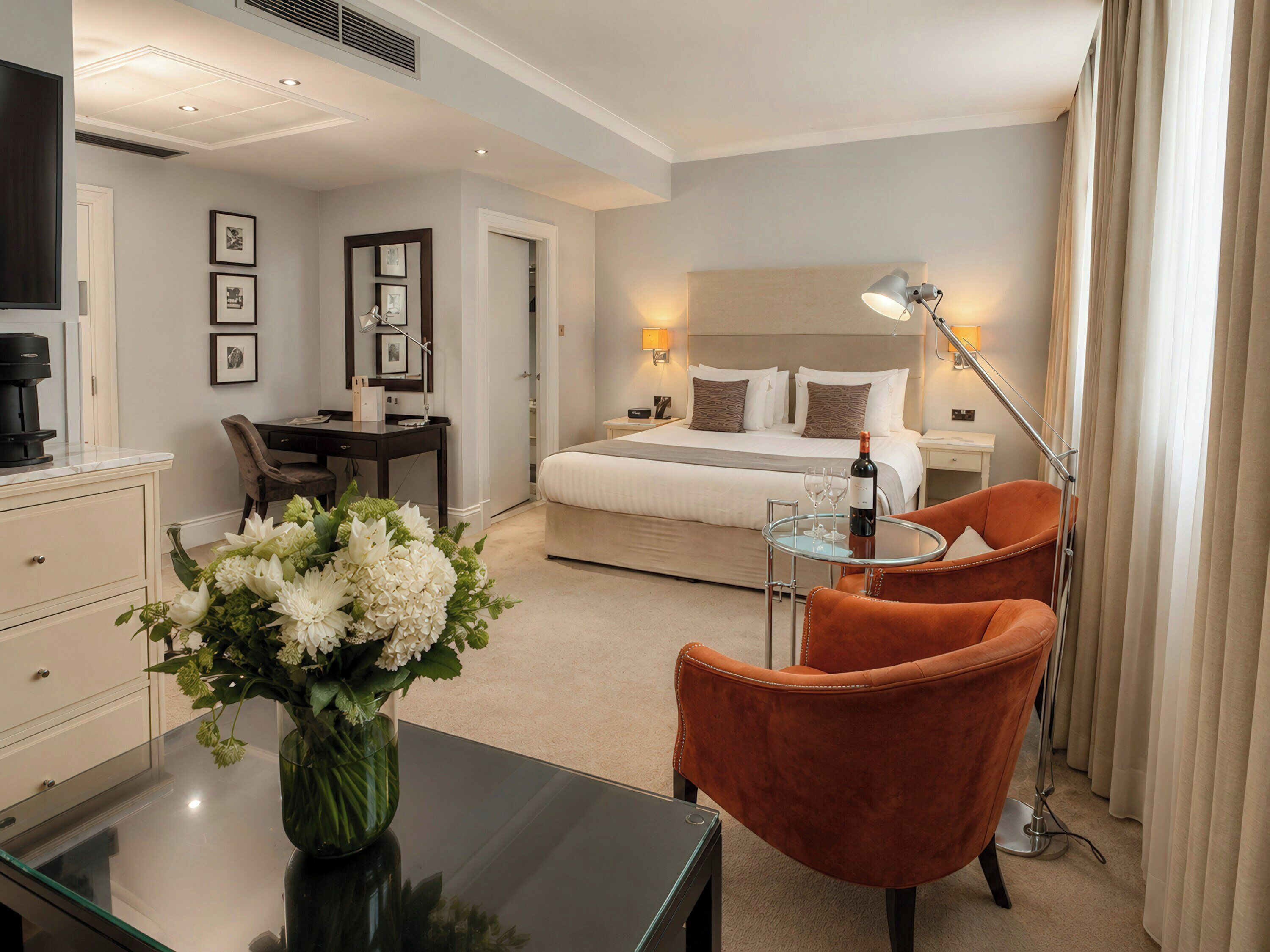 A hotel room with a bed, bedside table, chair, desk, and a vase of flowers.
