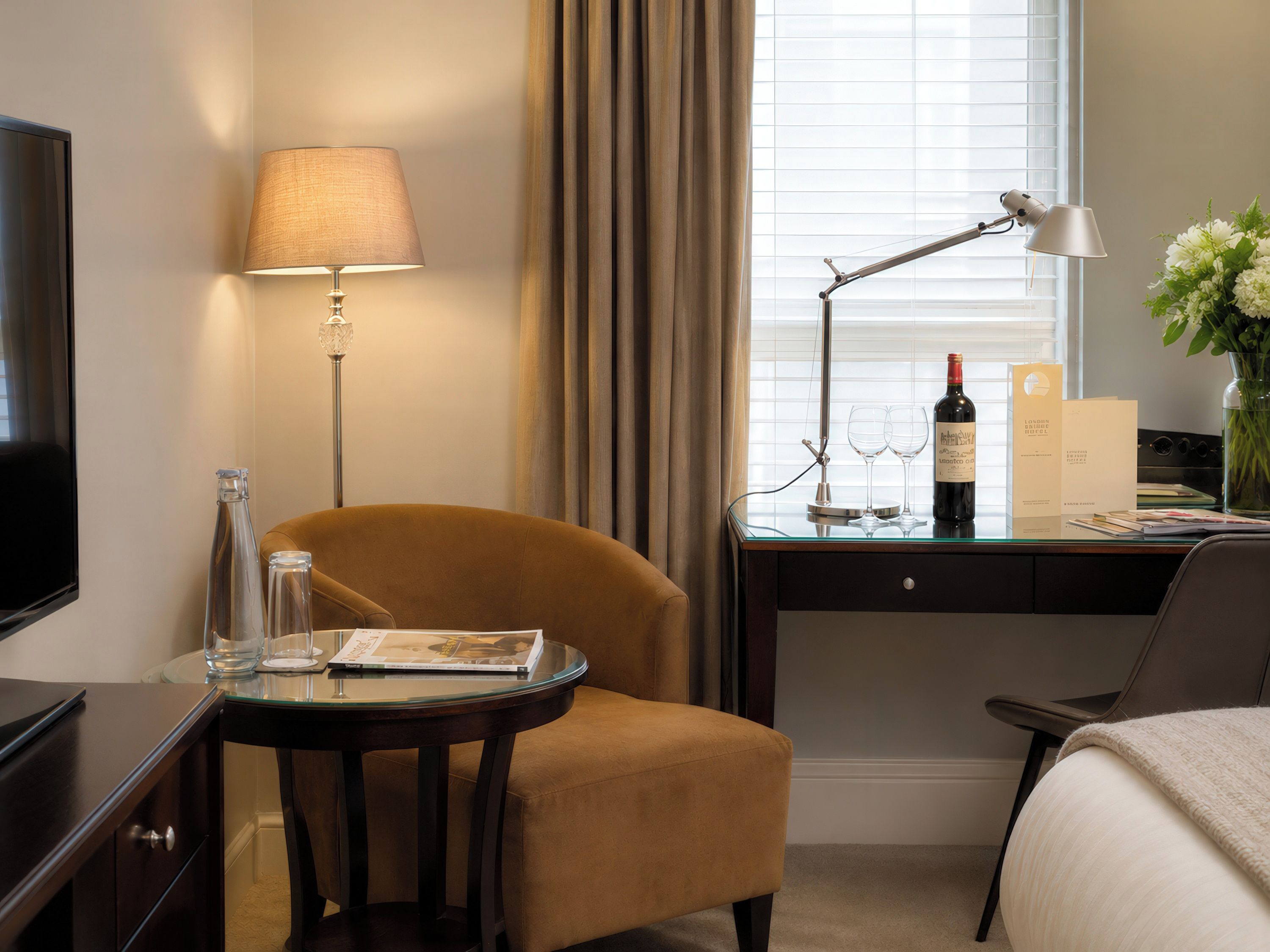 A hotel room with a bed, a desk with a lamp, a chair, a television, and a window with curtains.