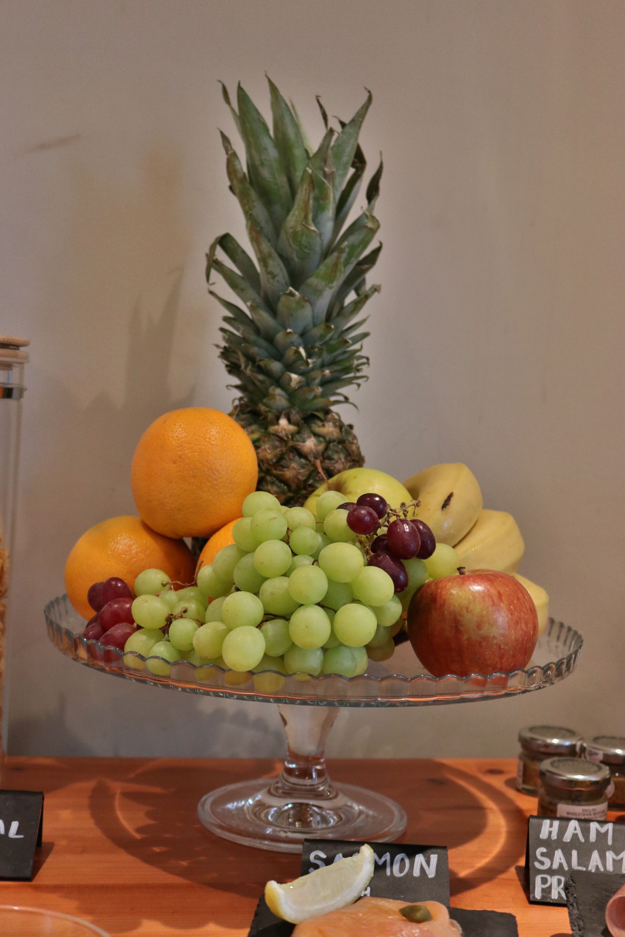 A fruit display on a glass stand with labels for ham, salmon, and prawn.