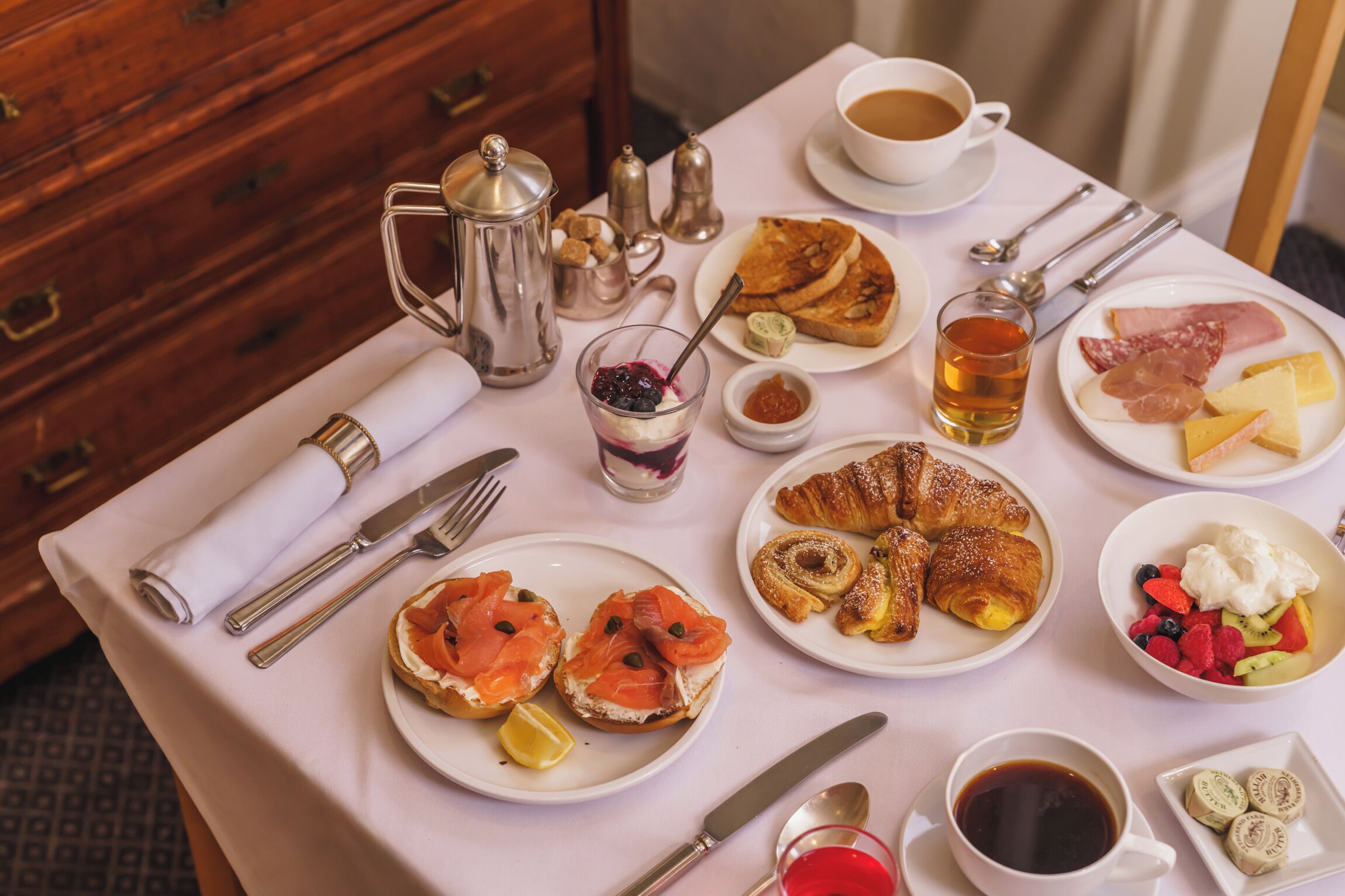 Une table de petit-déjeuner avec divers aliments, notamment des croissants, du saumon fumé et des fruits. On y trouve également des tasses de café et de thé, ainsi qu'une cafetière en argent.