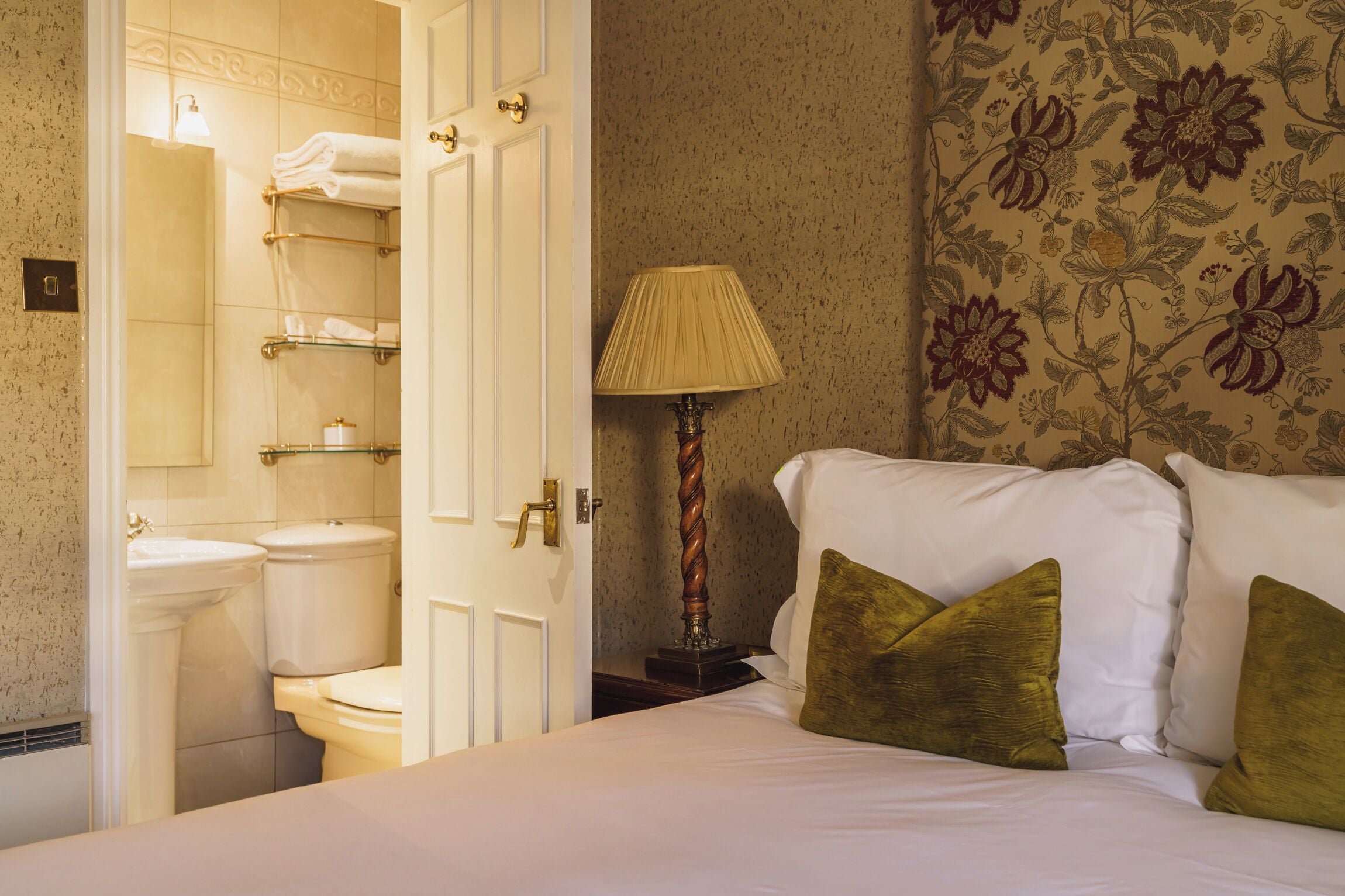 A hotel room with a bed, a nightstand, a lamp, a bathroom with a toilet and towel racks.