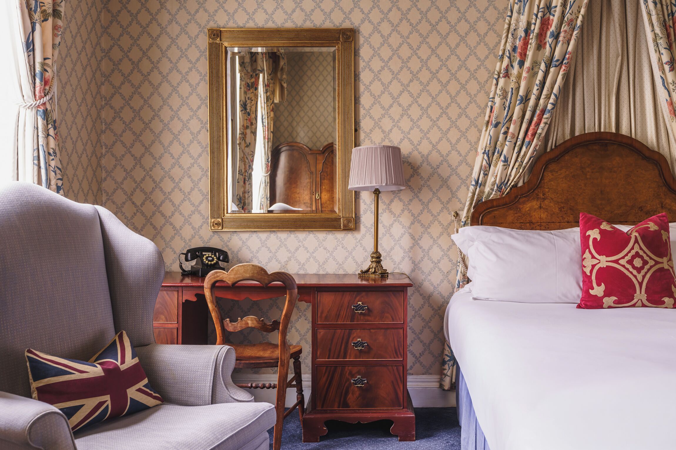 A hotel room with a wooden headboard, a bedside table with a lamp, a chair with a Union Jack pillow, and a bed with a red and gold pillow.
